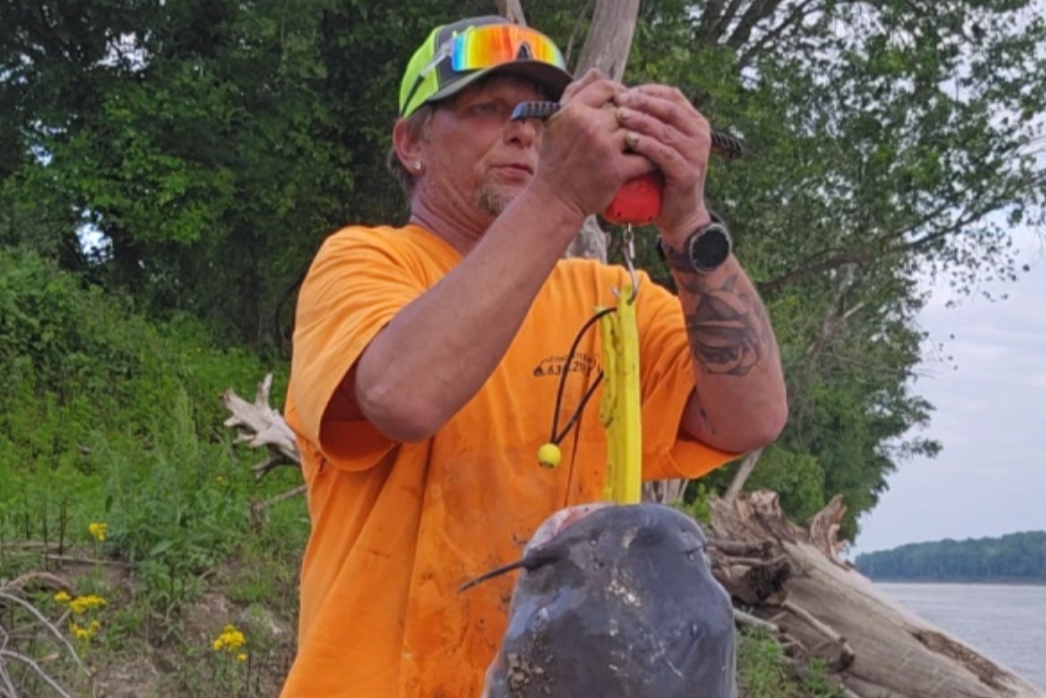 Person holding a large fish by a lakeside with trees and water in the background