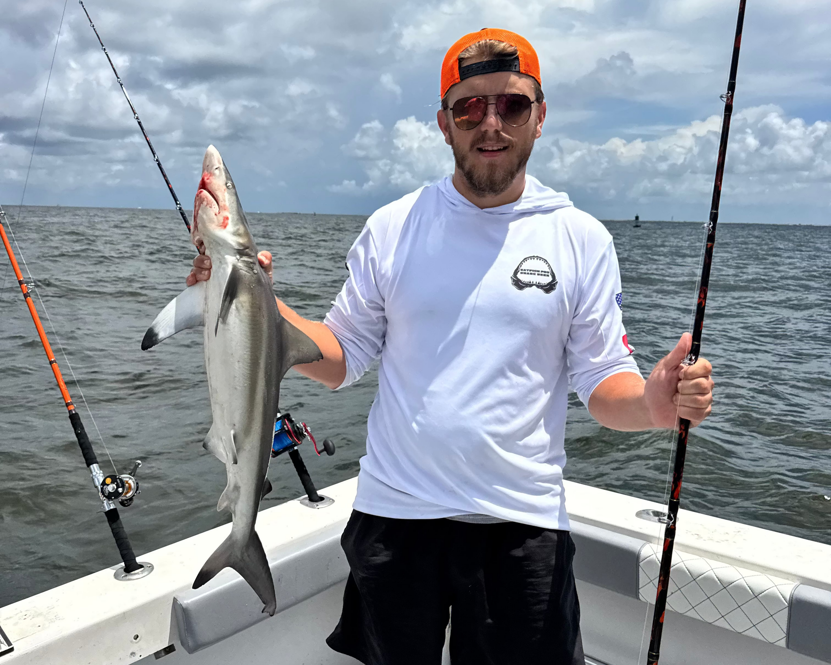 Man on a boat holding a fish with fishing rods, wearing a white shirt and black shorts, against a cloudy sky.