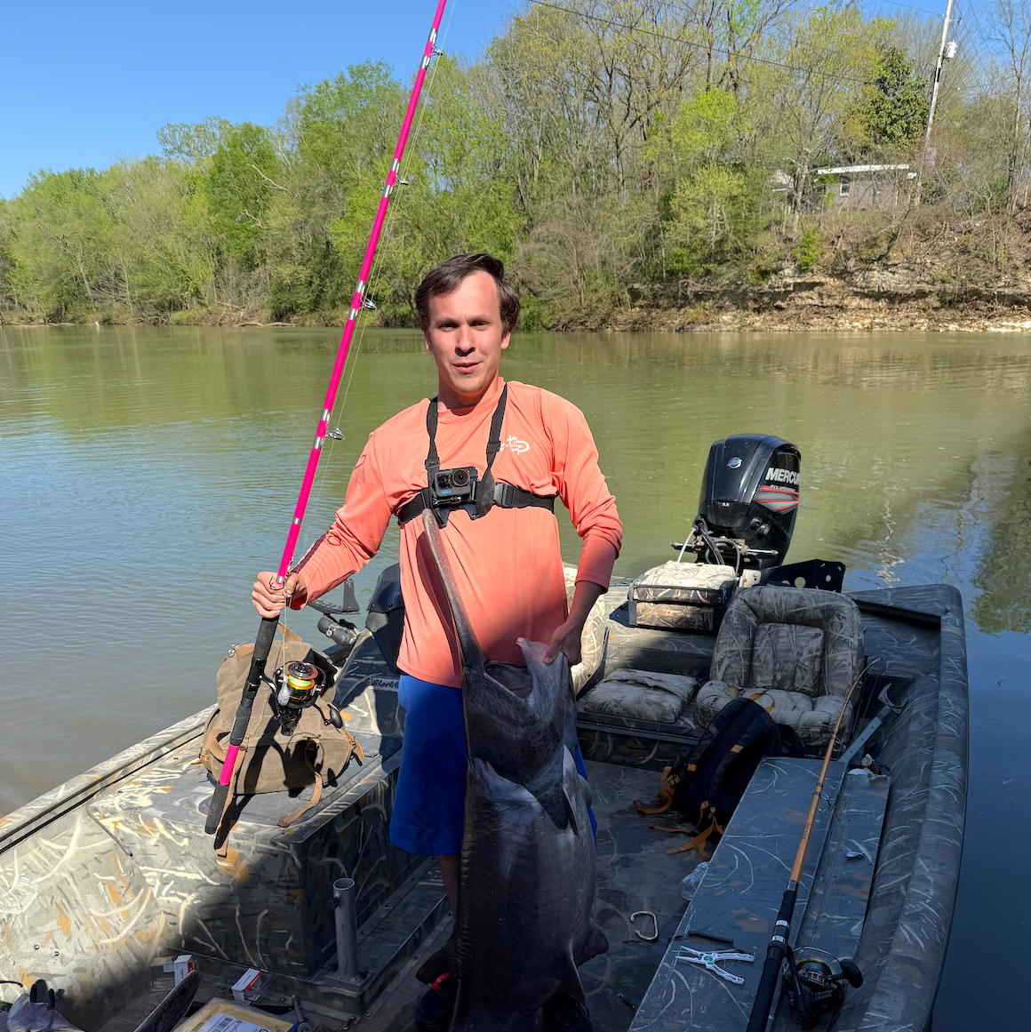 Man holding a large fish on a boat with fishing equipment on a lake.
